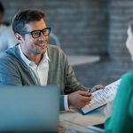 Feliz Empresario Conversando Com Seu Colega Enquanto Analisa Relatorios Financeiros No Escritorio 1 Scaled